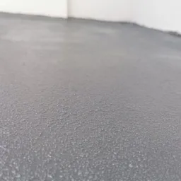 Szara, porowata powierzchnia z bliska, prawdopodobnie nowa izolacja przeciwwilgociowa fundamentów w narożniku pomieszczenia, widoczny fragment białej ściany.