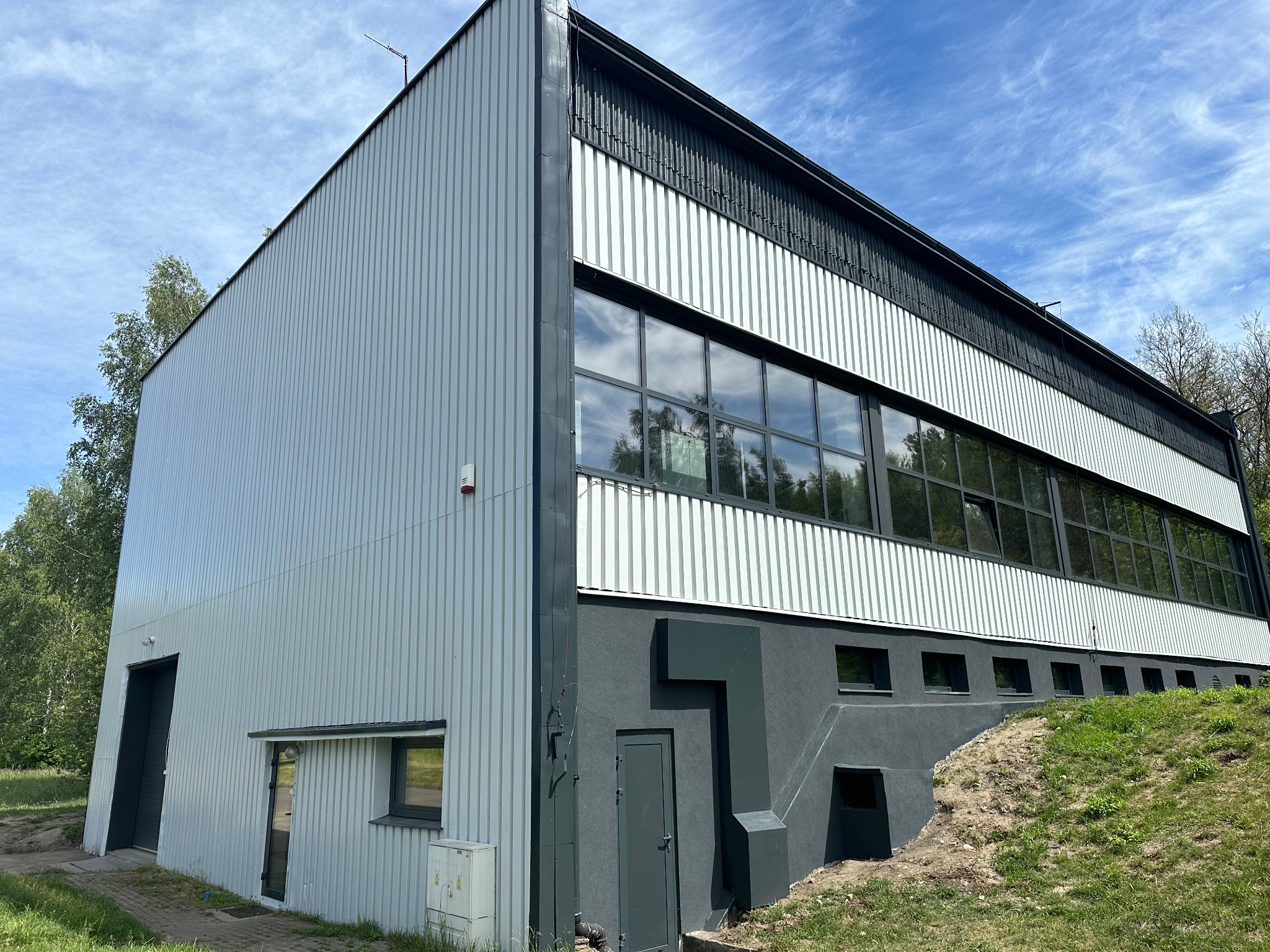 Nowoczesny budynek przemysłowy z szarej blachy trapezowej, z ciemnoszarą podmurówką i rzędem okien na górze, otoczony zielenią.