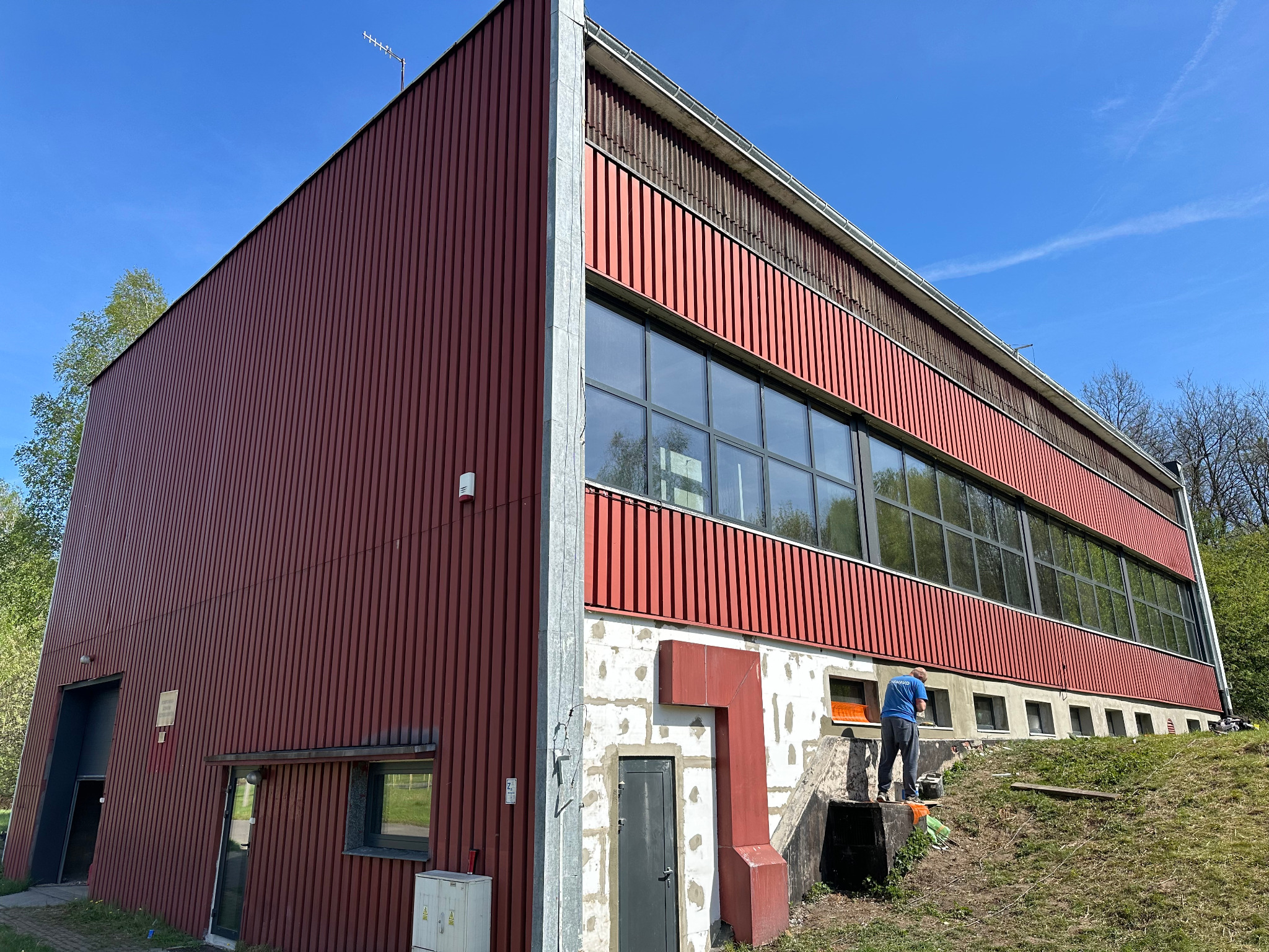 Budynek z elewacją w odcieniach czerwieni, z widocznymi oknami i fragmentem fundamentów w trakcie prac wykończeniowych, na zewnątrz pracownik w niebieskiej koszulce przy oknie.