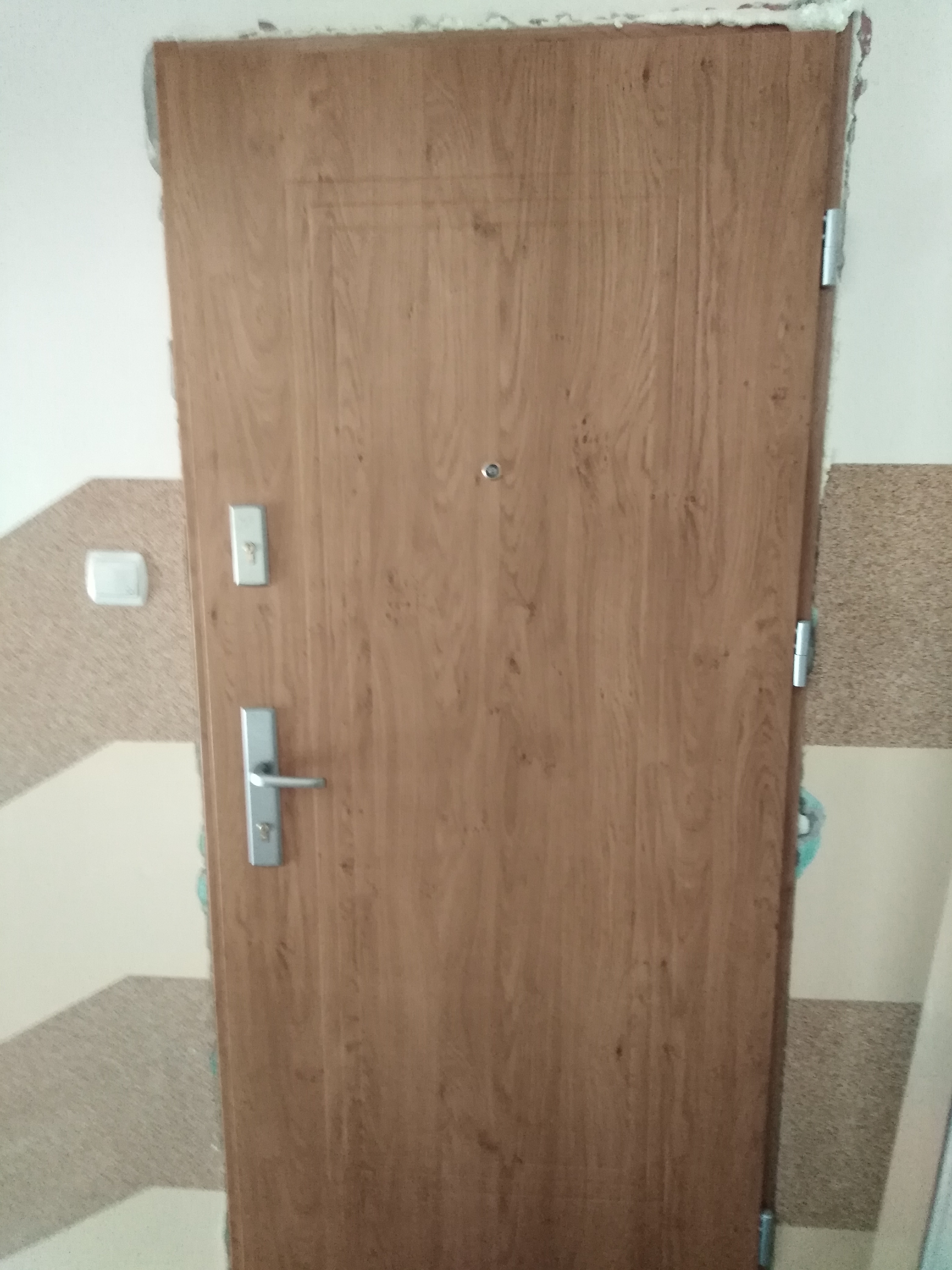 Drewniane drzwi wejściowe z wizjerem i zamkiem, widoczne ślady pianki montażowej wokół ościeżnicy, na ścianie obok włącznik światła.
