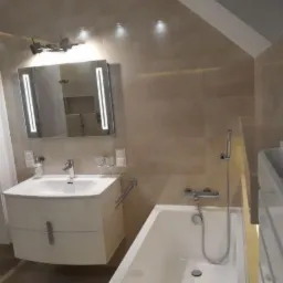 Jasna łazienka na poddaszu z wanną, umywalką z szafką i podświetlanym lustrem, wykończona beżowymi płytkami.