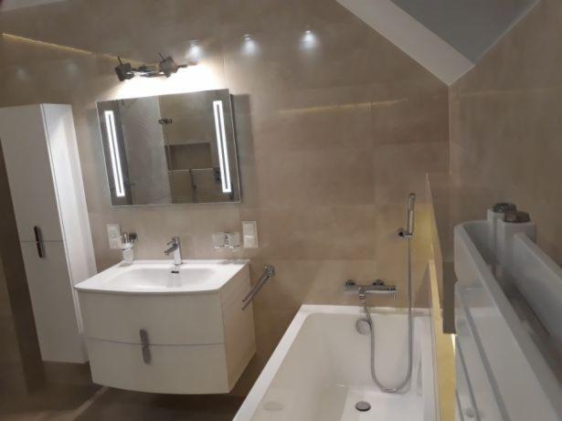 Jasna łazienka na poddaszu z wanną, umywalką z szafką i podświetlanym lustrem, wykończona beżowymi płytkami.