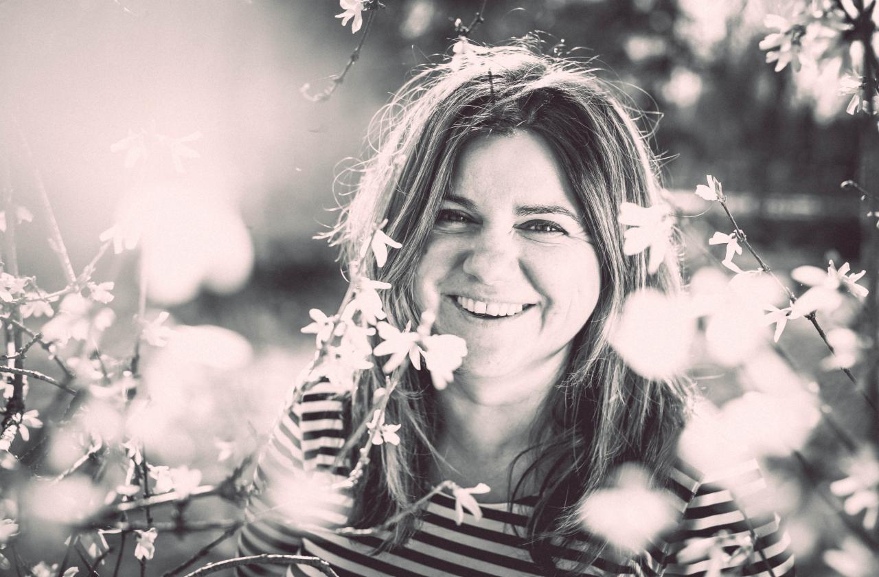 Uśmiechnięta kobieta w paski otoczona kwitnącymi gałązkami, portret w sepii z delikatnym rozmyciem tła i promieniami słońca.