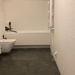 NOWUS-BUD Sp. z o.o. - Nowoczesna łazienka z białymi płytkami imitującymi kamień, ciemną podłogą, wiszącą toaletą i czarną armaturą, widoczny grzejnik drabinkowy.