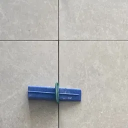 Szare płytki z niebieskim klipsem poziomującym i zielonym paskiem, ukazujące precyzję układania. Widok z bliska na łączenie płytek.