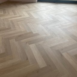 NOWUS-BUD Sp. z o.o. - Jasne panele ułożone w jodełkę francuską, świeżo po montażu. Widoczne delikatne różnice w odcieniach drewna i subtelne światło wpadające przez okno.