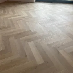 Jasne panele ułożone w jodełkę francuską, świeżo po montażu. Widoczne delikatne różnice w odcieniach drewna i subtelne światło wpadające przez okno.