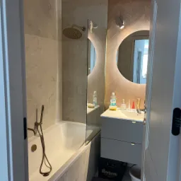 Beżowa łazienka z wanną, szklanym parawanem, okrągłym lustrem z podświetleniem i szarą szafką pod umywalką. Na ścianach płytki imitujące kamień, na jednej ze ścian drewniana mozaika.