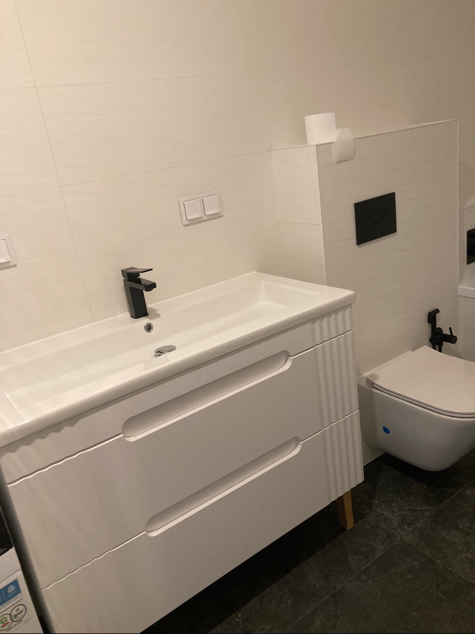 Nowoczesna łazienka z białą umywalką z szafką na drewnianych nóżkach, czarną armaturą i podwieszaną toaletą z bidetką na tle białych płytek.