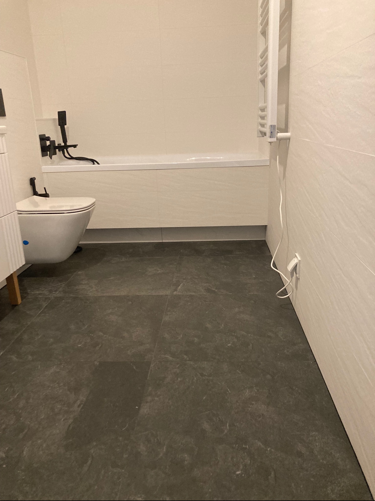 Nowoczesna łazienka z wiszącą toaletą, wanną z czarną armaturą i grzejnikiem drabinkowym, wykończona płytkami w odcieniach bieli i szarości, z ciemną podłogą imitującą kamień.