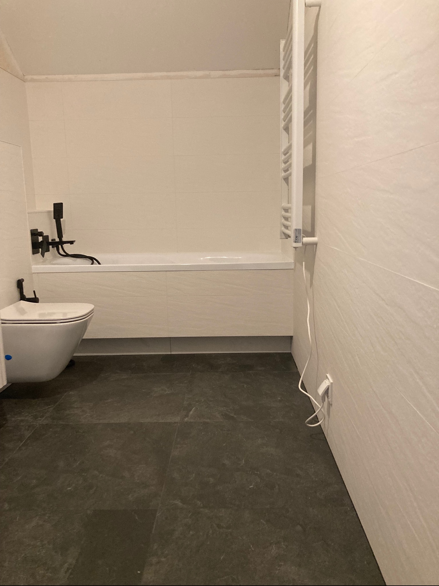 Nowoczesna łazienka z białymi płytkami imitującymi kamień, ciemną podłogą, wiszącą toaletą i czarną armaturą, widoczny grzejnik drabinkowy.