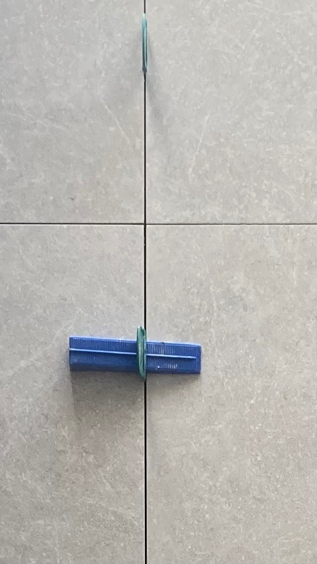 Szare płytki z niebieskim klipsem poziomującym i zielonym paskiem, ukazujące precyzję układania. Widok z bliska na łączenie płytek.