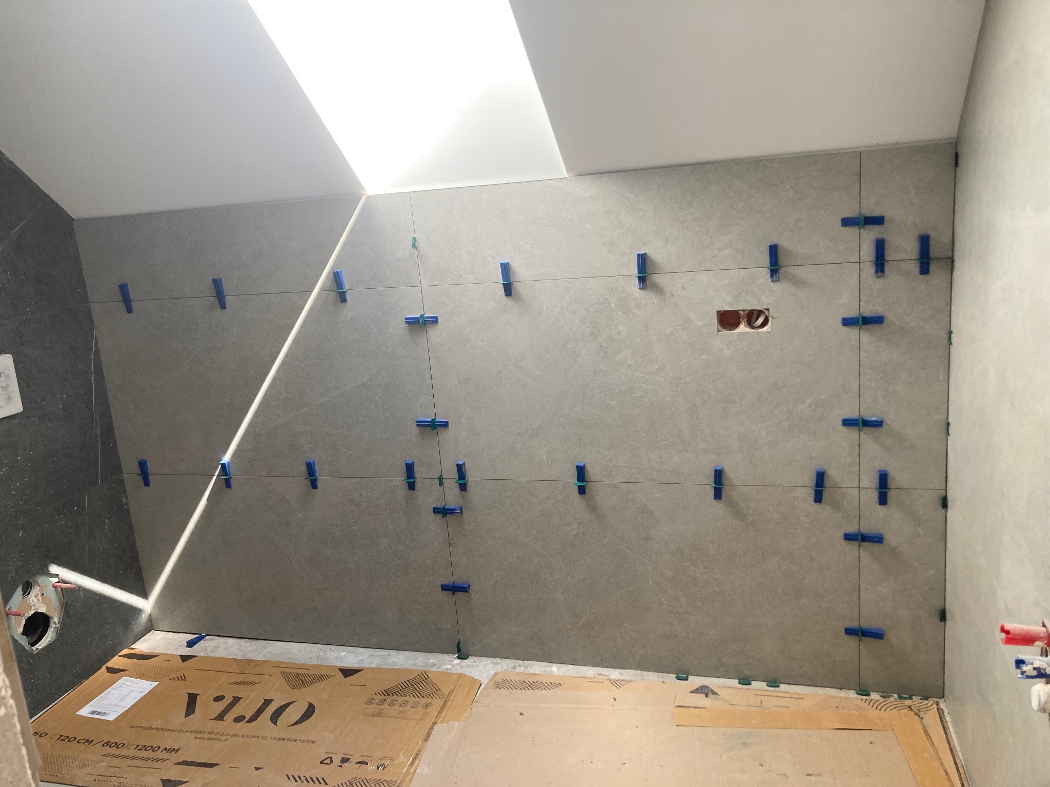 Ściana łazienki w trakcie układania szarych płytek wielkoformatowych, z użyciem niebieskich klipsów poziomujących i sznurka. Karton po płytkach na podłodze.