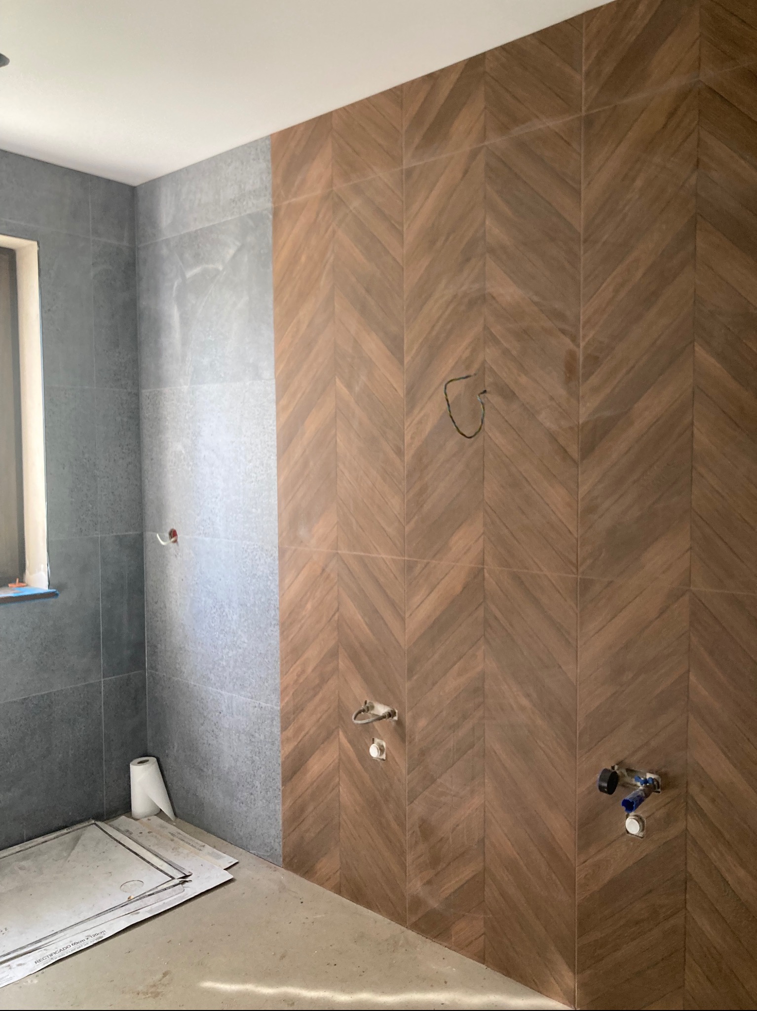 Łazienka w trakcie remontu: szare płytki na ścianie obok okna i brązowe płytki ułożone w jodełkę na sąsiedniej ścianie. Widoczne instalacje wod-kan.