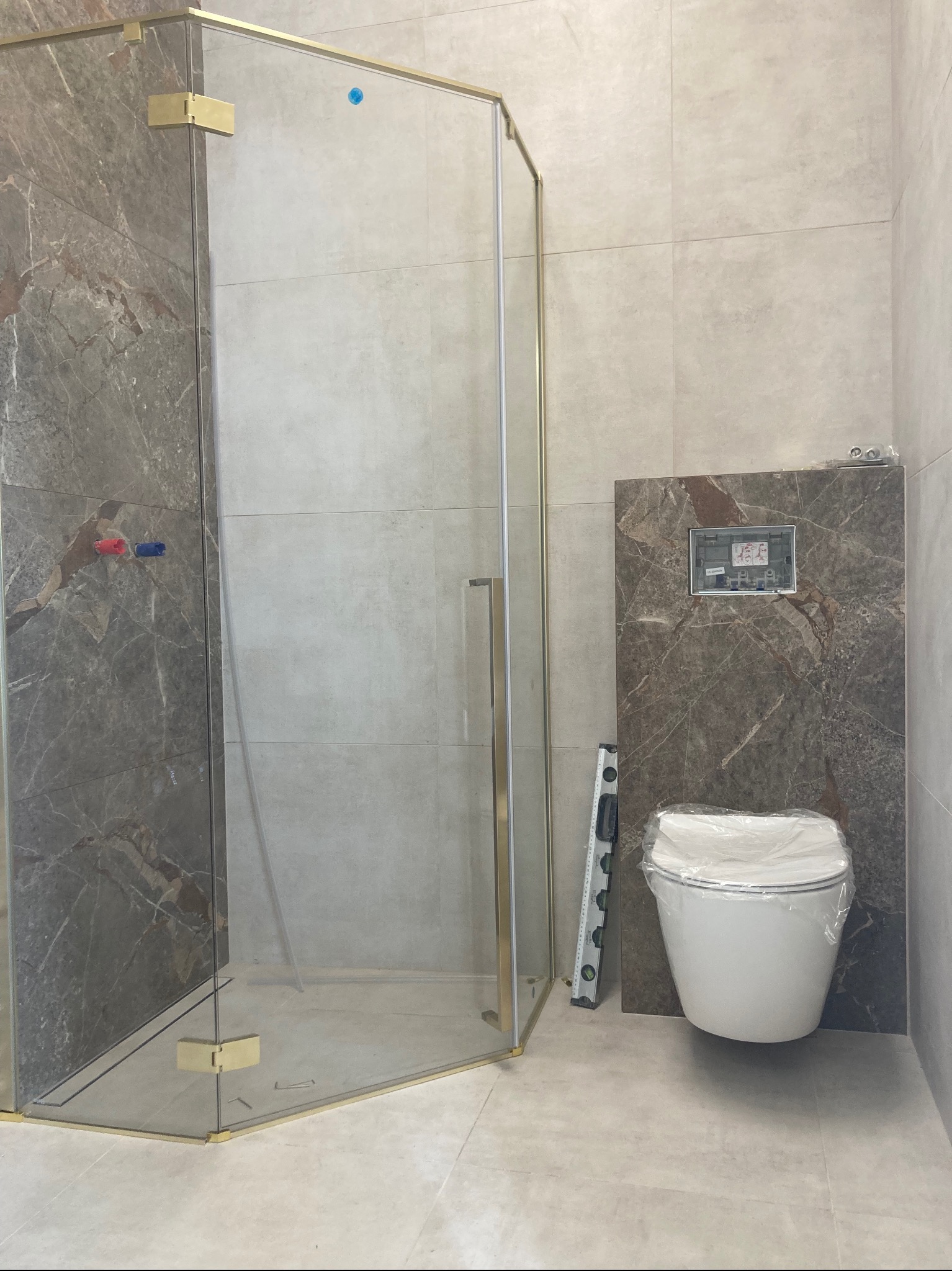 Nowoczesna łazienka w trakcie wykańczania: kabina prysznicowa ze złotymi okuciami, podwieszana toaleta z poziomicą i marmurowe płytki.