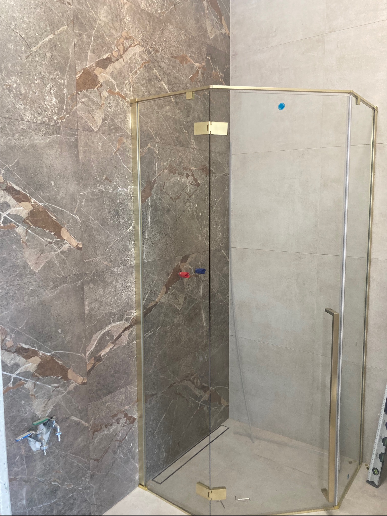 Narożna kabina prysznicowa z złotymi profilami na tle płytek imitujących marmur i gładkich, jasnych płytek. Widoczne elementy montażowe i poziomica.