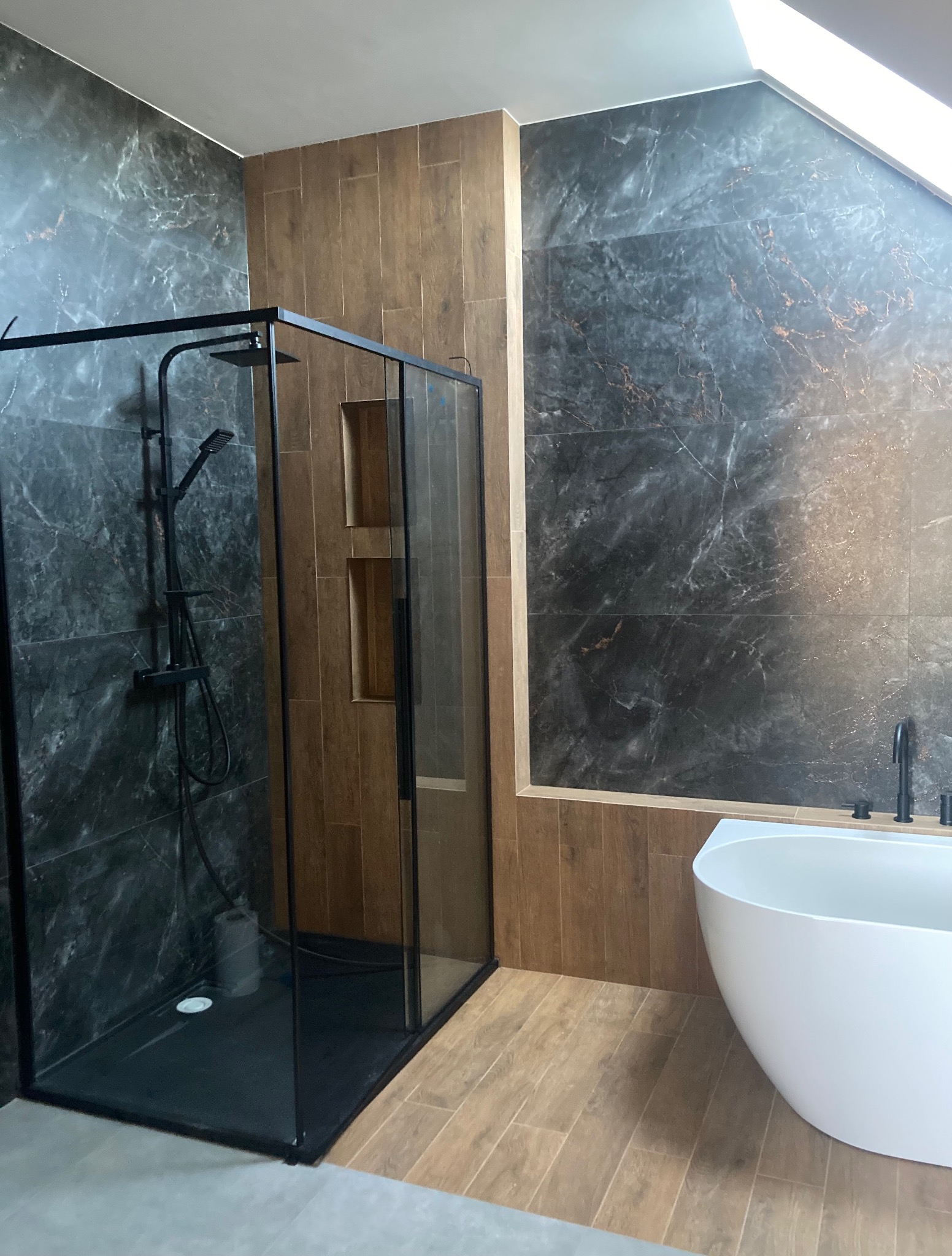 Nowoczesna łazienka z kabiną typu walk-in z czarnymi profilami, płytkami imitującymi marmur i drewno oraz wolnostojącą białą wanną. Widok na skośny sufit.