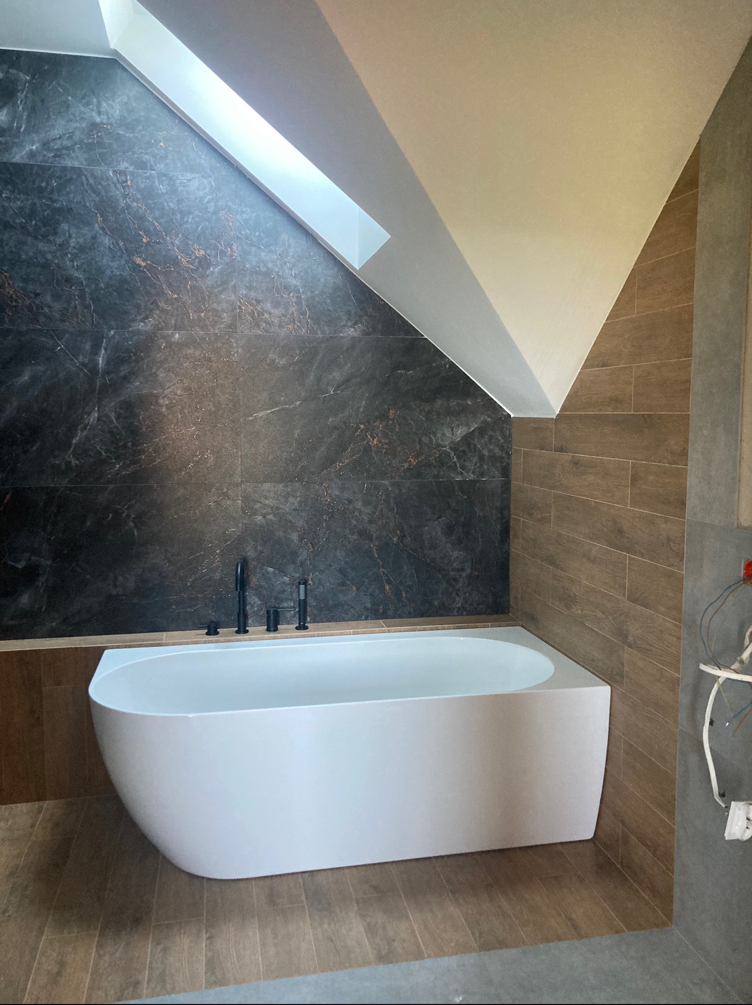 Nowoczesna wanna wolnostojąca w łazience na poddaszu z marmurową ścianą i drewnopodobnymi płytkami. Uwagę zwraca czarna armatura i skos.