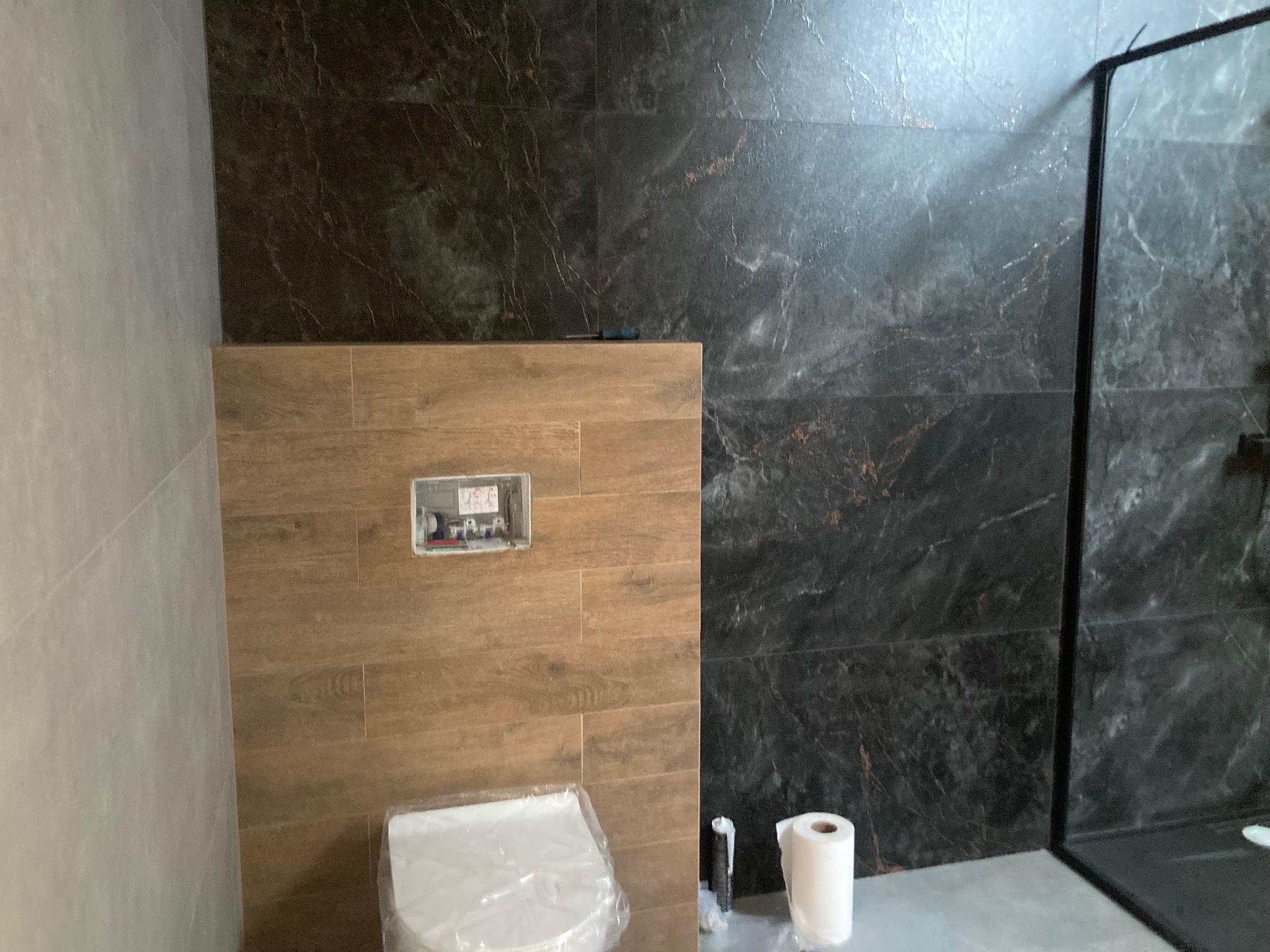 Nowoczesna łazienka z marmurowymi płytkami i drewnopodobną zabudową WC. Widoczny fragment kabiny prysznicowej i rolka papieru toaletowego.