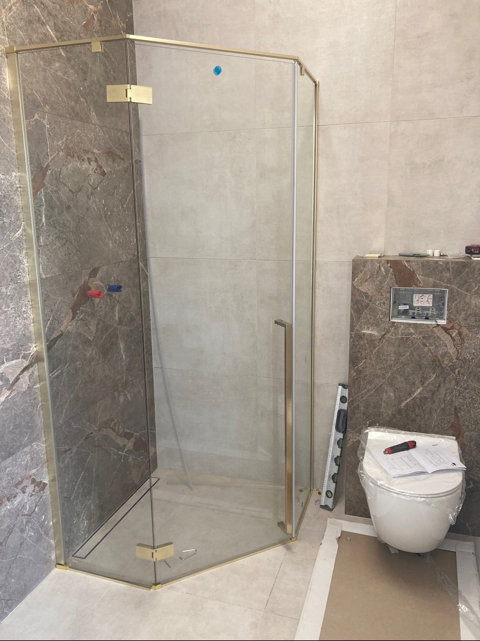 Narożna kabina prysznicowa z złotymi profilami, zamontowana w łazience z płytkami imitującymi marmur. Widoczny fragment toalety i poziomica.