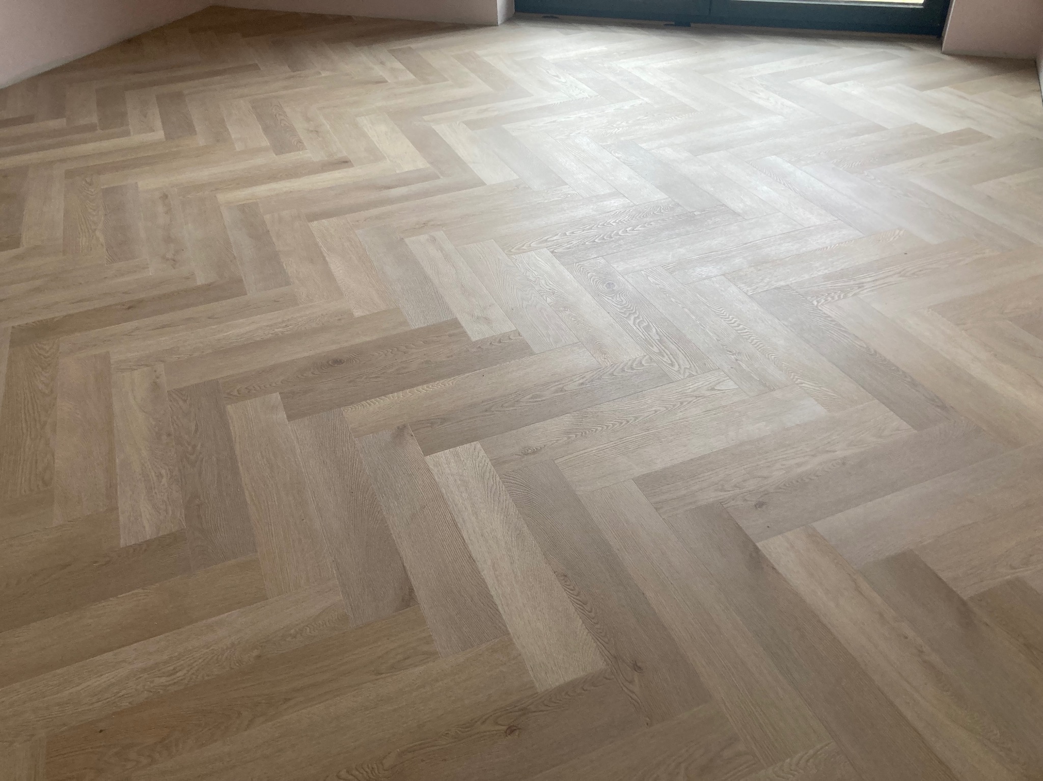 Jasne panele ułożone w jodełkę francuską, świeżo po montażu. Widoczne delikatne różnice w odcieniach drewna i subtelne światło wpadające przez okno.