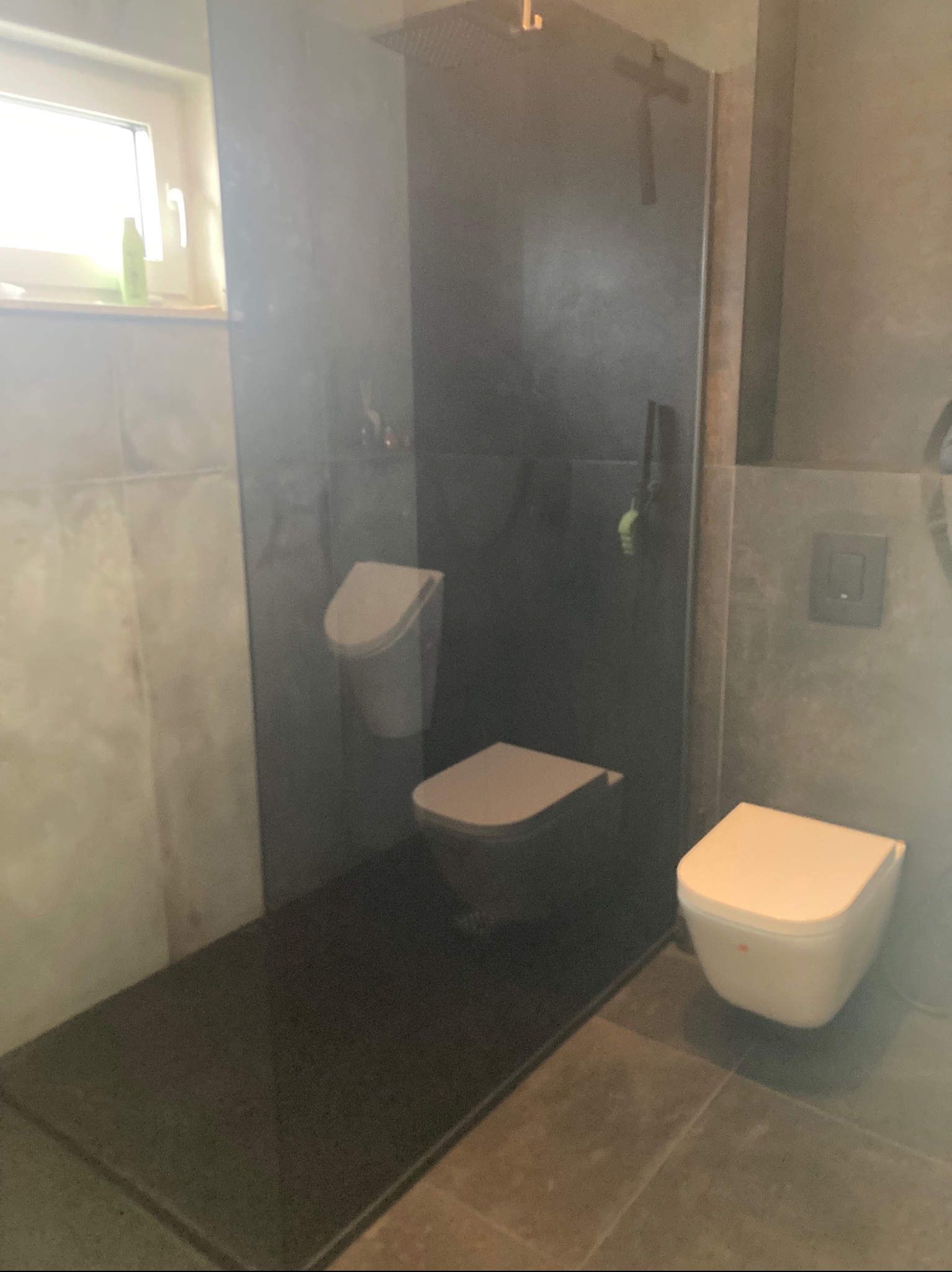 Nowoczesna łazienka z kabiną typu walk-in, szarą armaturą i płytkami w odcieniach szarości. Minimalistyczny wystrój, widoczny fragment okna z rośliną.