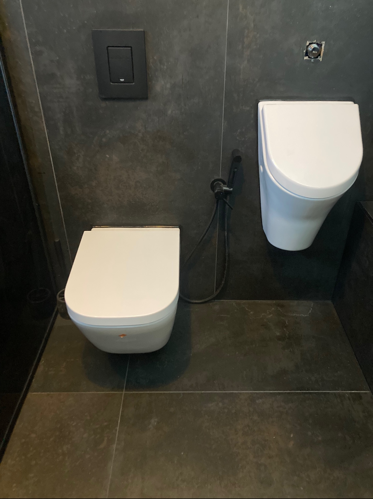 Nowoczesna toaleta podwieszana z bidetką i czarnym przyciskiem na ścianie z szarych płytek. Minimalistyczny design łazienki w ciemnej kolorystyce.