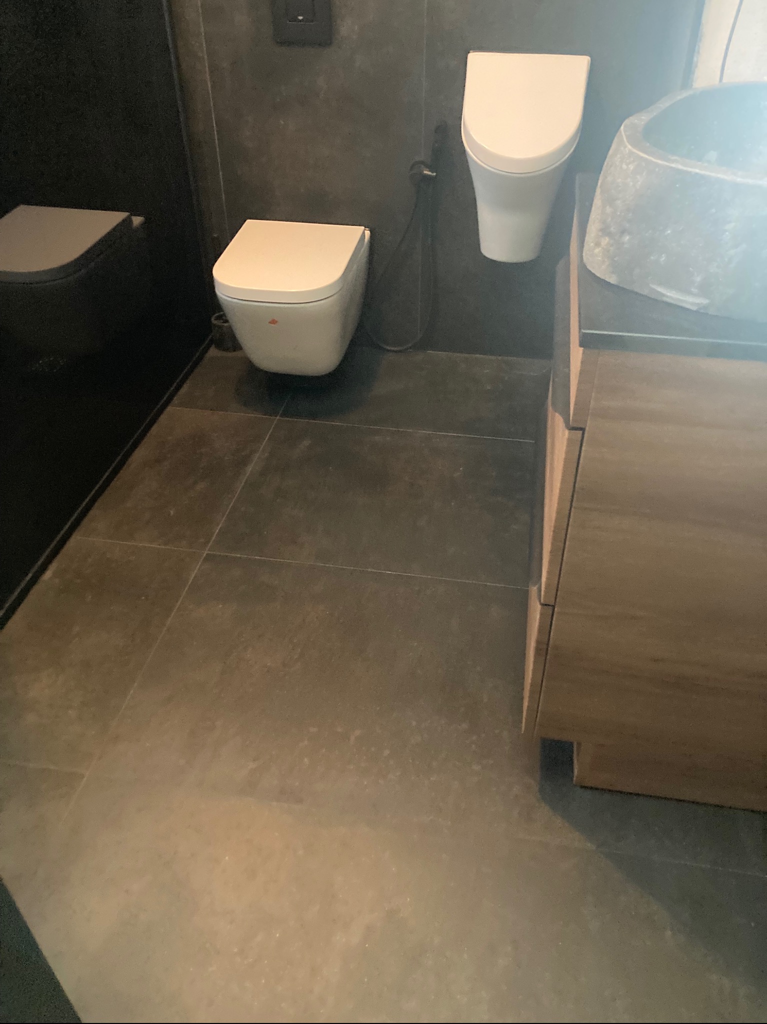 Nowoczesna łazienka z szarą armaturą i kamienną umywalką. Widoczne podwieszane WC i płytki podłogowe w odcieniach szarości. Minimalistyczny design.