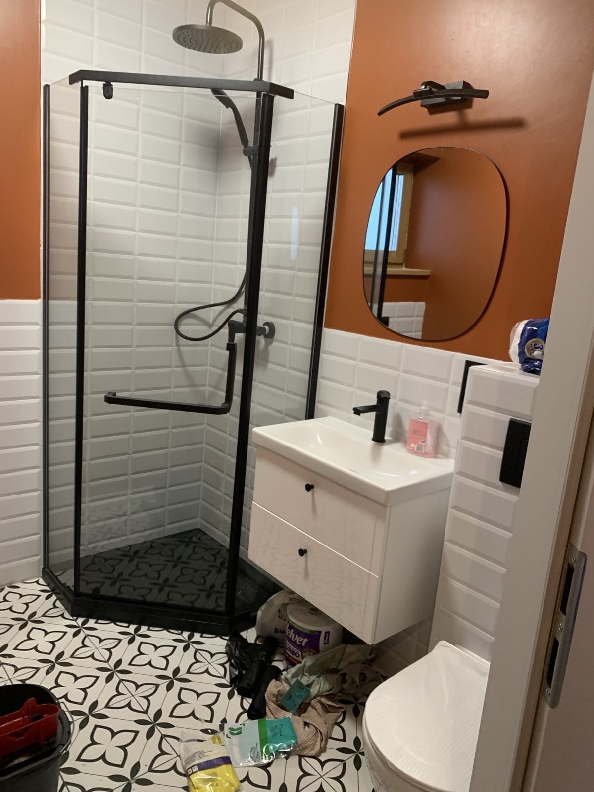 Nowoczesna łazienka po remoncie: kabina prysznicowa z czarną armaturą, biała umywalka z szafką, lustro o nieregularnym kształcie na ścianie w kolorze terakoty.