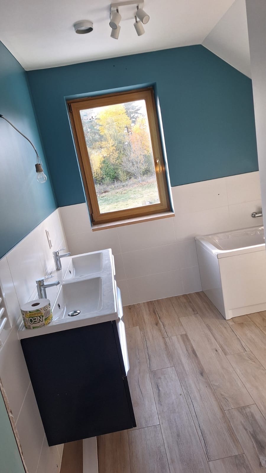 Wykończona łazienka na poddaszu z drewnopodobną podłogą, podwójną umywalką i oknem z widokiem na jesienne drzewa. Ściany w odcieniach bieli i turkusu.