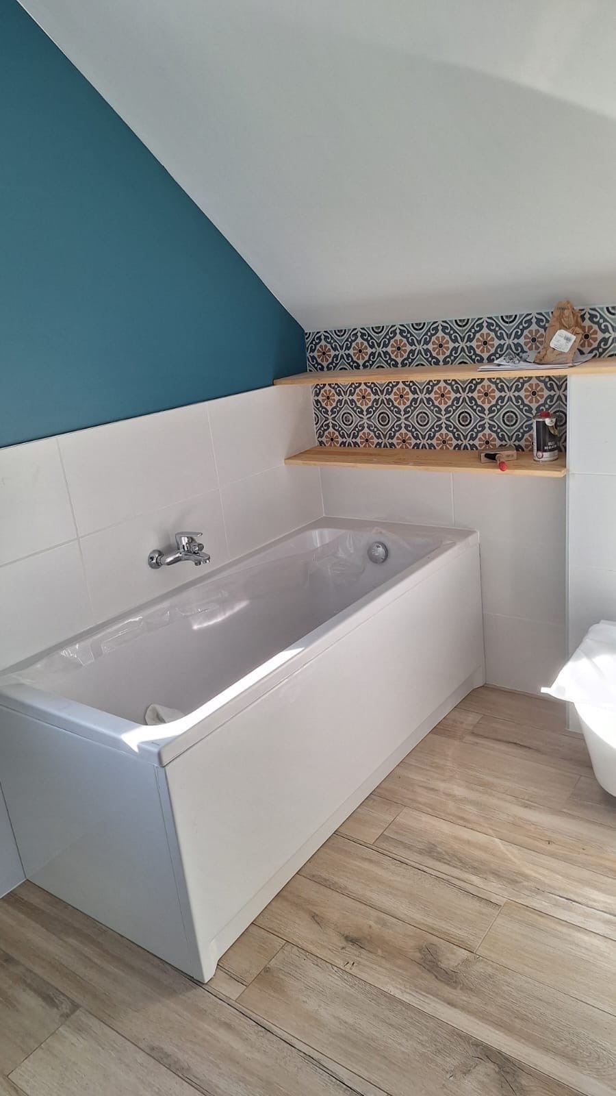 Biała wanna w łazience na poddaszu z drewnopodobną podłogą. Niebieska ściana, półka z dekoracyjnymi płytkami i akcesoriami. Ukończony remont, widok na wannę i fragment toalety.