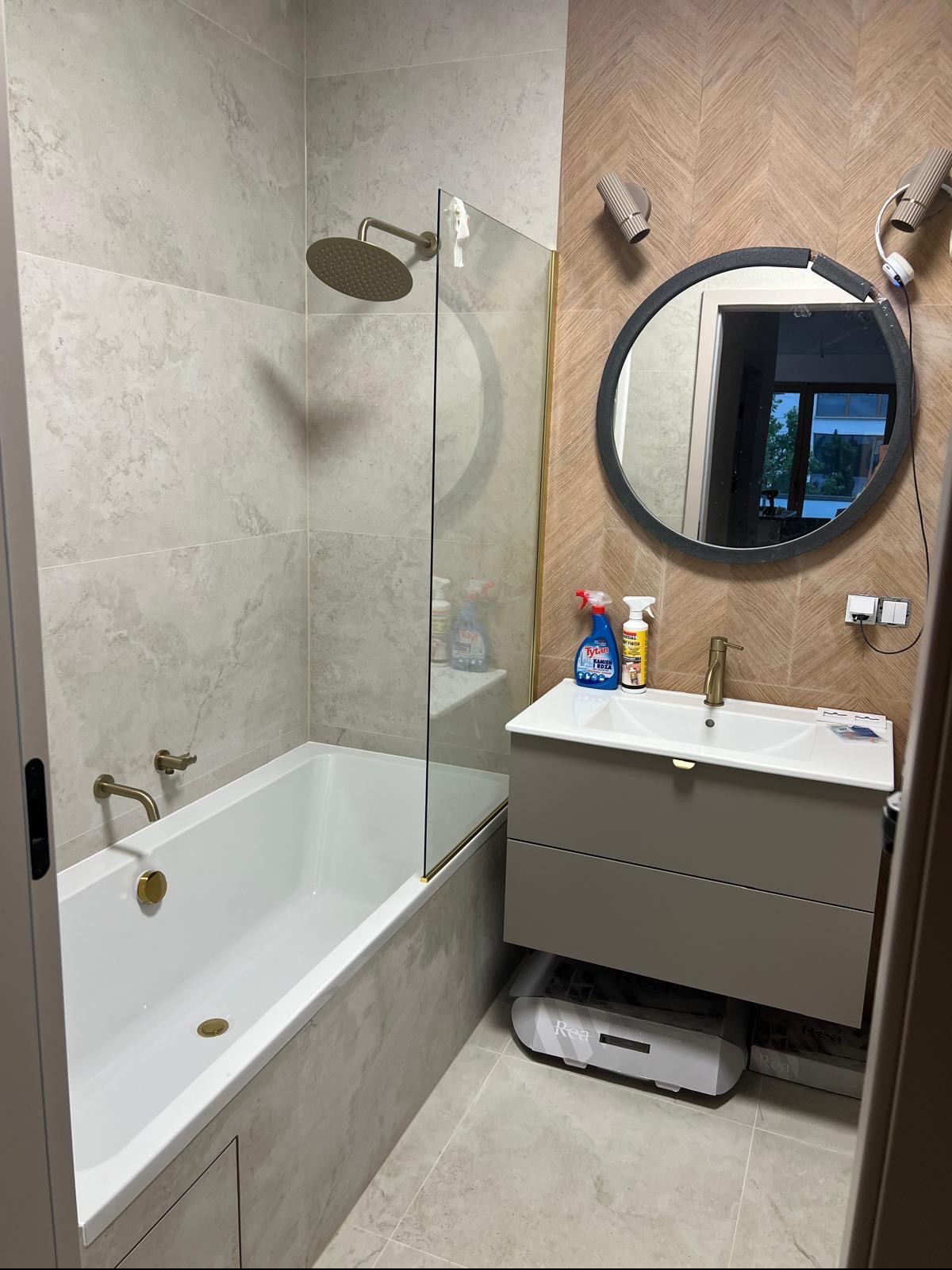 Wykończona łazienka z wanną, przeszklonym parawanem, umywalką z szafką w odcieniach szarości i okrągłym lustrem na ścianie z wzorem jodełki.