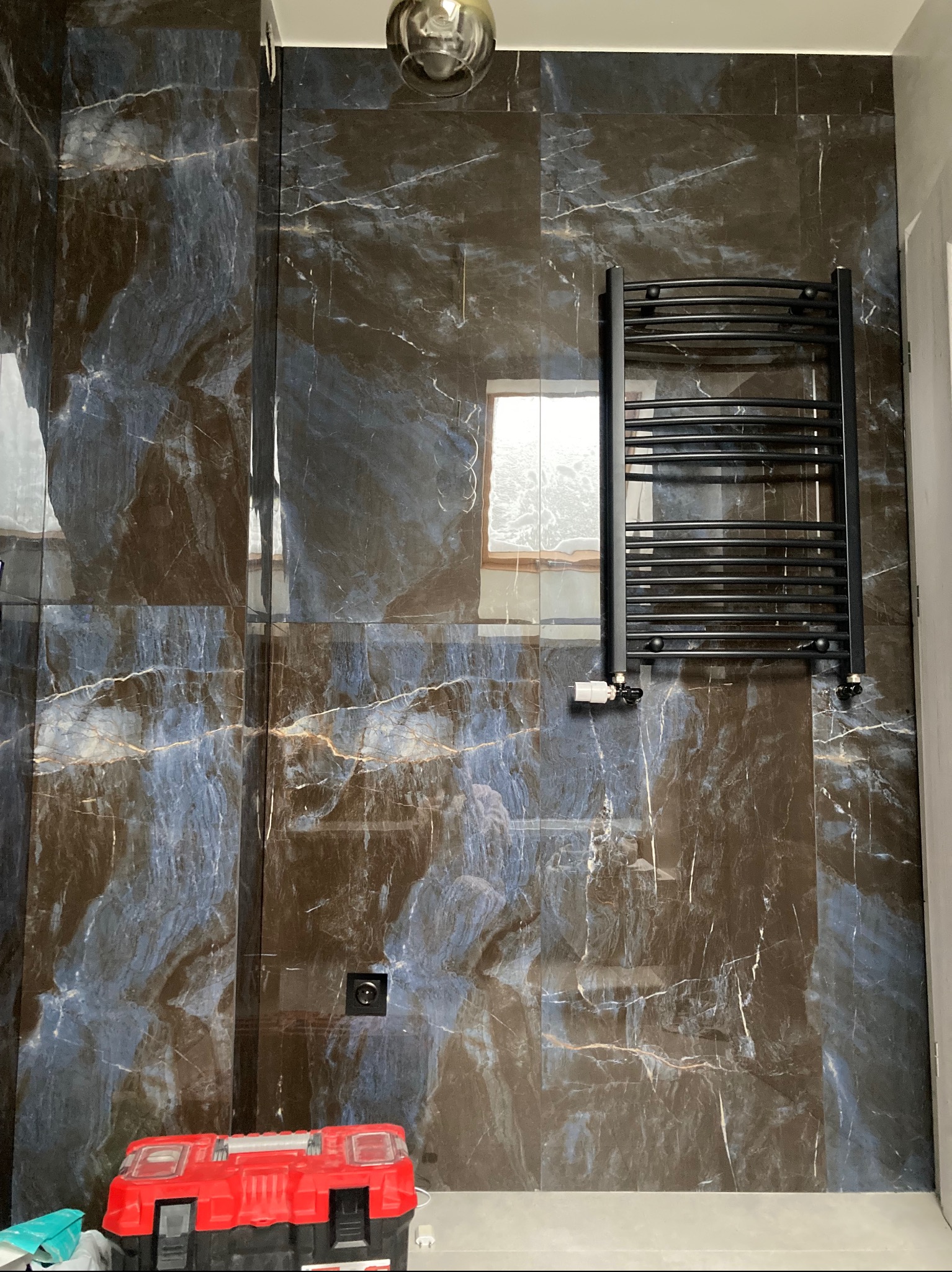 Ciemne, marmurowe płytki w łazience z zamontowanym grzejnikiem drabinkowym. Widoczna skrzynka z narzędziami i gniazdko elektryczne.
