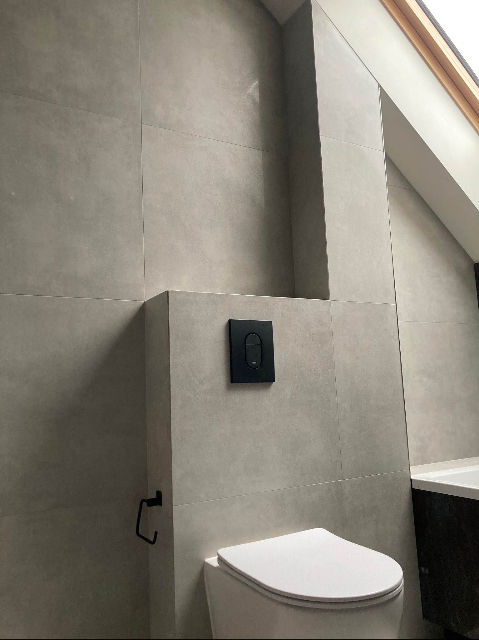 Wykończona łazienka na poddaszu z szarymi, matowymi płytkami, podwieszaną toaletą i czarnymi akcentami. Widoczne okno dachowe i fragment wanny.