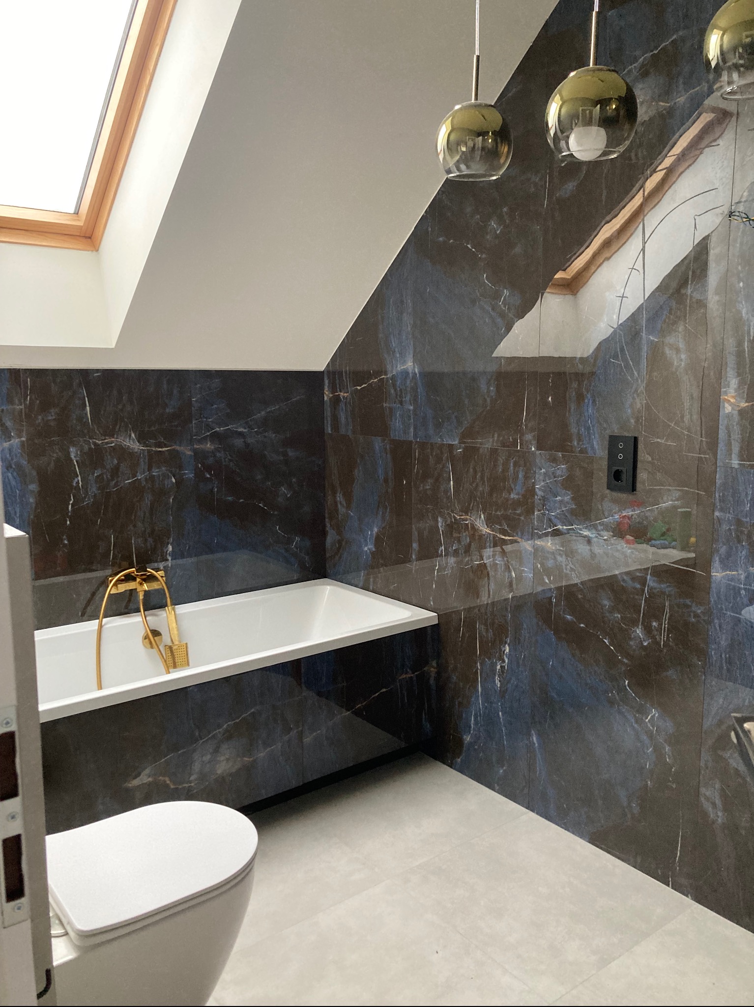 Stylowa łazienka na poddaszu z oknem dachowym. Ciemne płytki imitujące marmur, złota armatura przy białej wannie i wiszące lampy w stylu glamour.