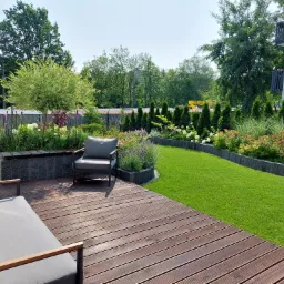 Nowoczesny ogród z drewnianym tarasem, szarymi meblami ogrodowymi i starannie utrzymanym trawnikiem, obok budynku mieszkalnego.
