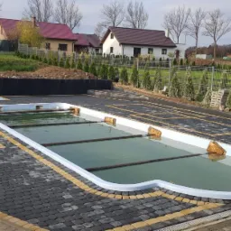 Nowo wybudowany staw kąpielowy z szarą kostką brukową i żółtymi akcentami, otoczony tujami i ogrodzeniem z siatki, w tle domy jednorodzinne.