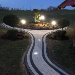 Oświetlona ścieżka z kostki brukowej w odcieniach szarości, prowadząca do okrągłego klombu z drzewami i lampami, widok po zmierzchu.