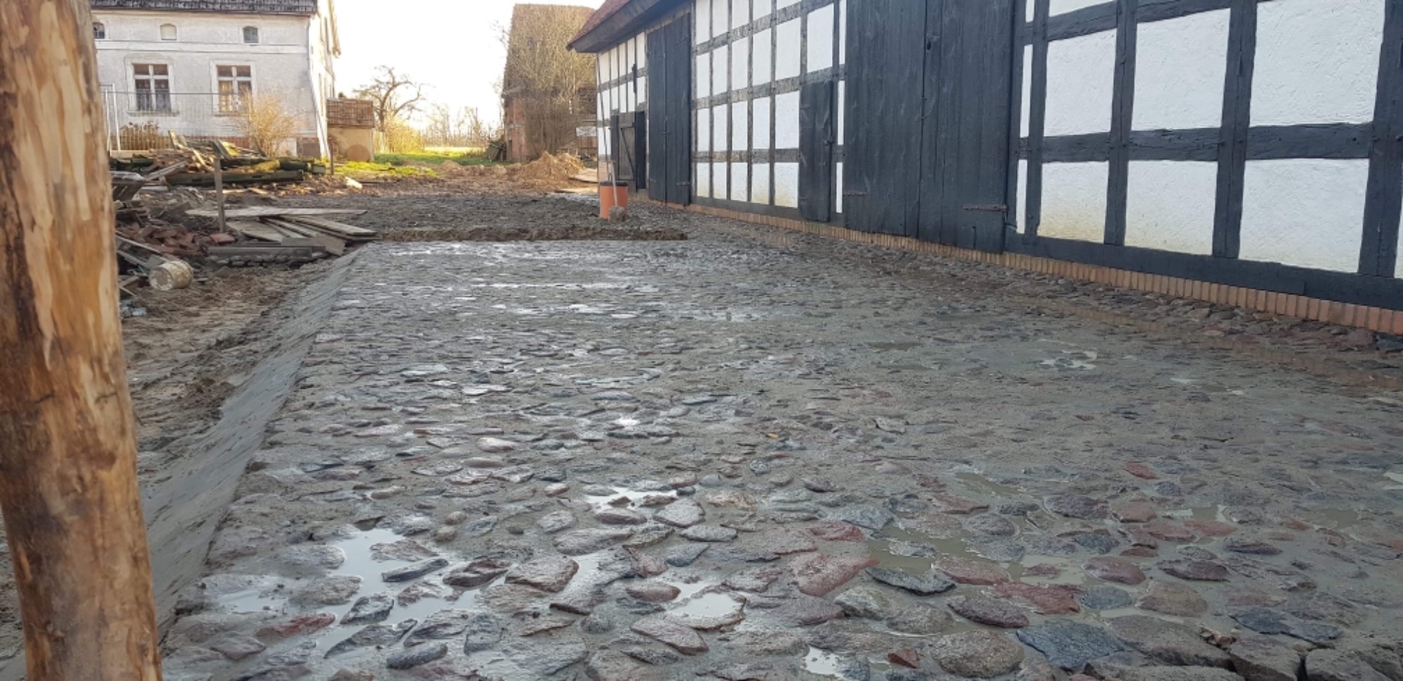 Ułożona, mokra kostka granitowa na podwórzu starego domu z muru pruskiego, widoczne pozostałości po budowie i drewniany słupek w kadrze.