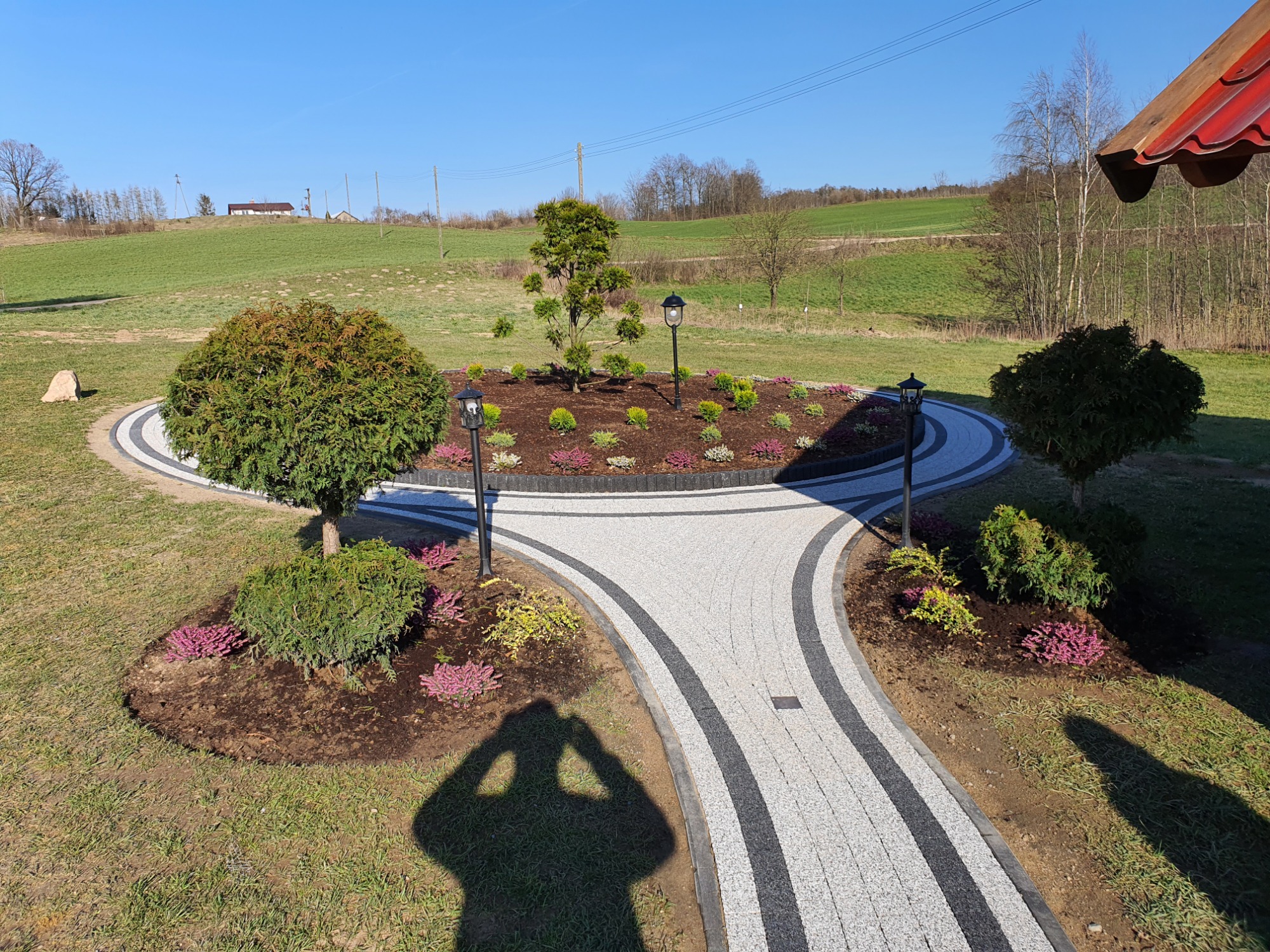 Ogród z okrągłą ścieżką z kostki brukowej w kolorze szarym i czarnym, rabaty z krzewami i kwiatami, oświetlone lampami ogrodowymi, na tle zielonego krajobrazu i błękitnego nieba. Cień osoby...