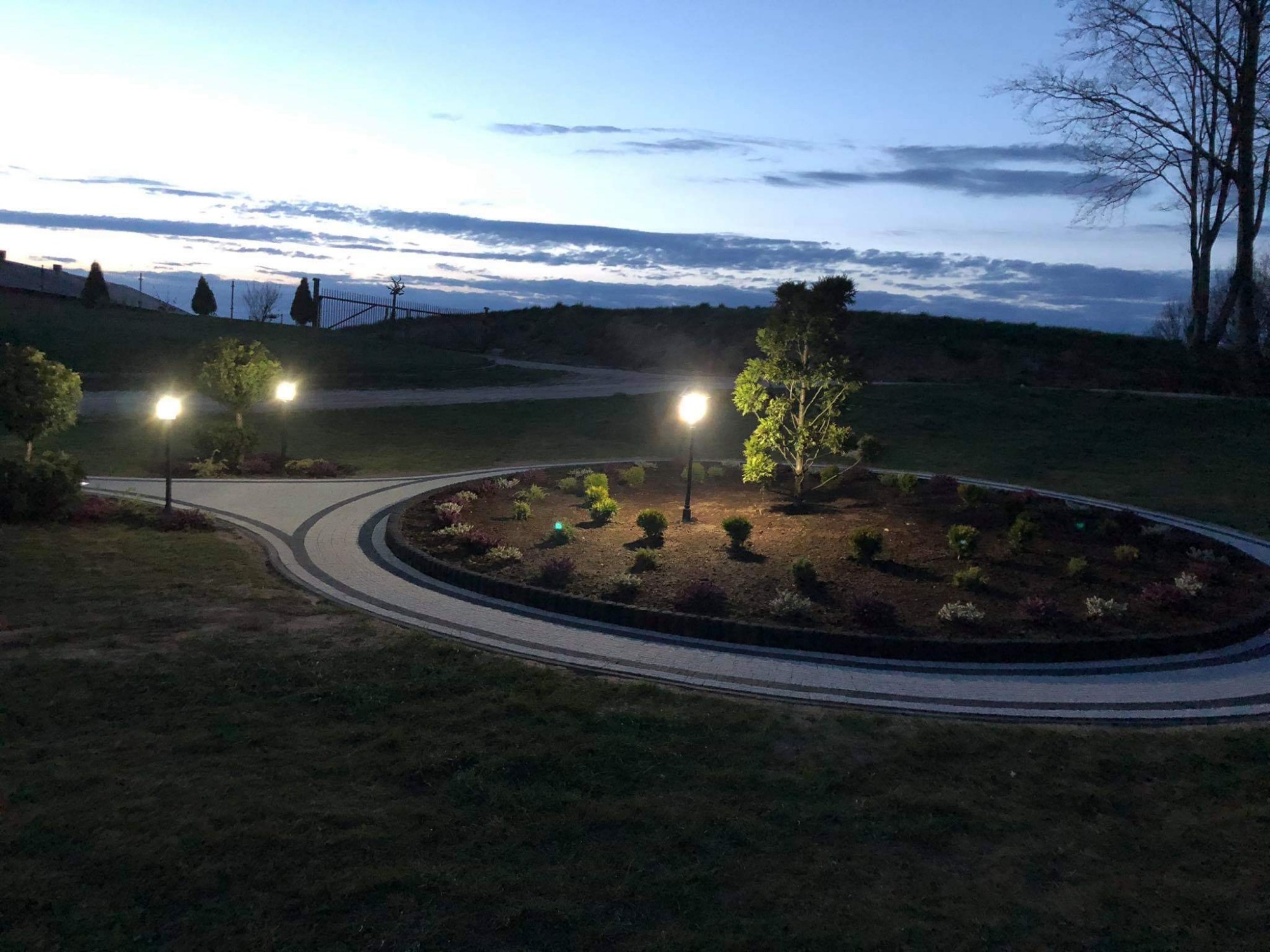 Oświetlony, okrągły klomb z nasadzeniami i ścieżką z jasnej kostki w Gowidlinie, widok o zmierzchu.