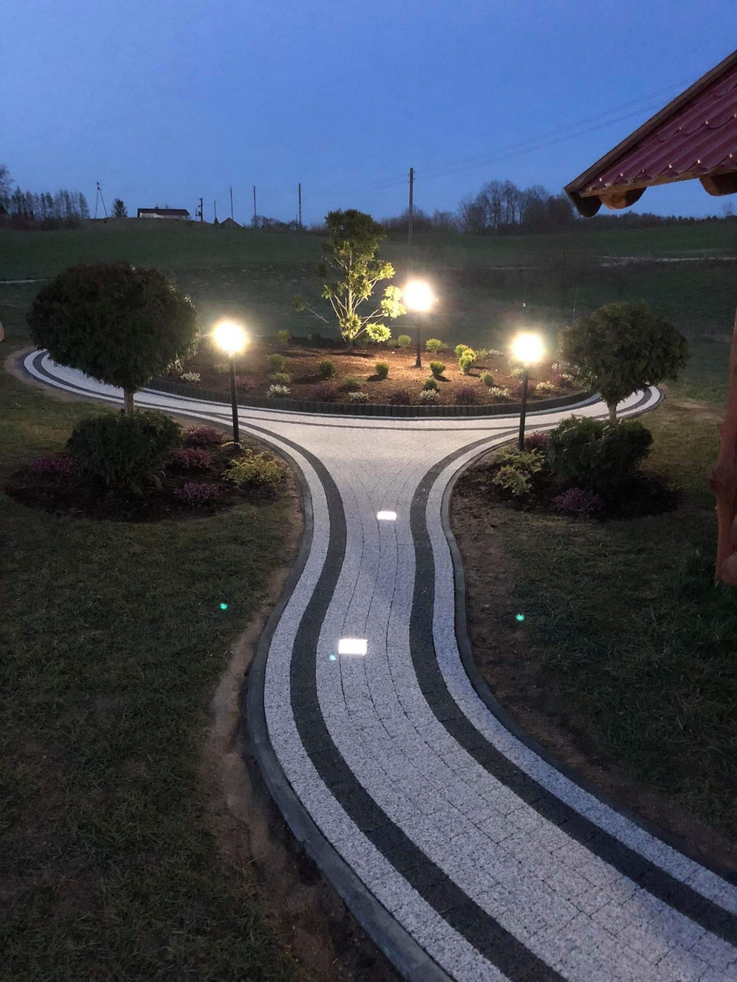 Oświetlona ścieżka z kostki brukowej w odcieniach szarości, prowadząca do okrągłego klombu z drzewami i lampami, widok po zmierzchu.