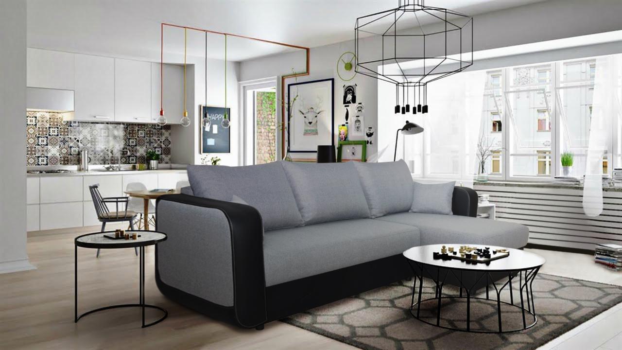 Szara sofa narożna z czarnymi elementami w nowoczesnym salonie z kuchnią w tle; na stoliku szachownica.