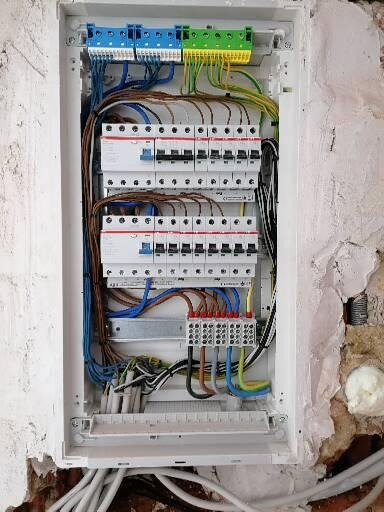 Otwarta skrzynka elektryczna z zainstalowanymi bezpiecznikami i przewodami różnej grubości oraz koloru, zamontowana na nierównej ścianie z widocznymi śladami tynku.