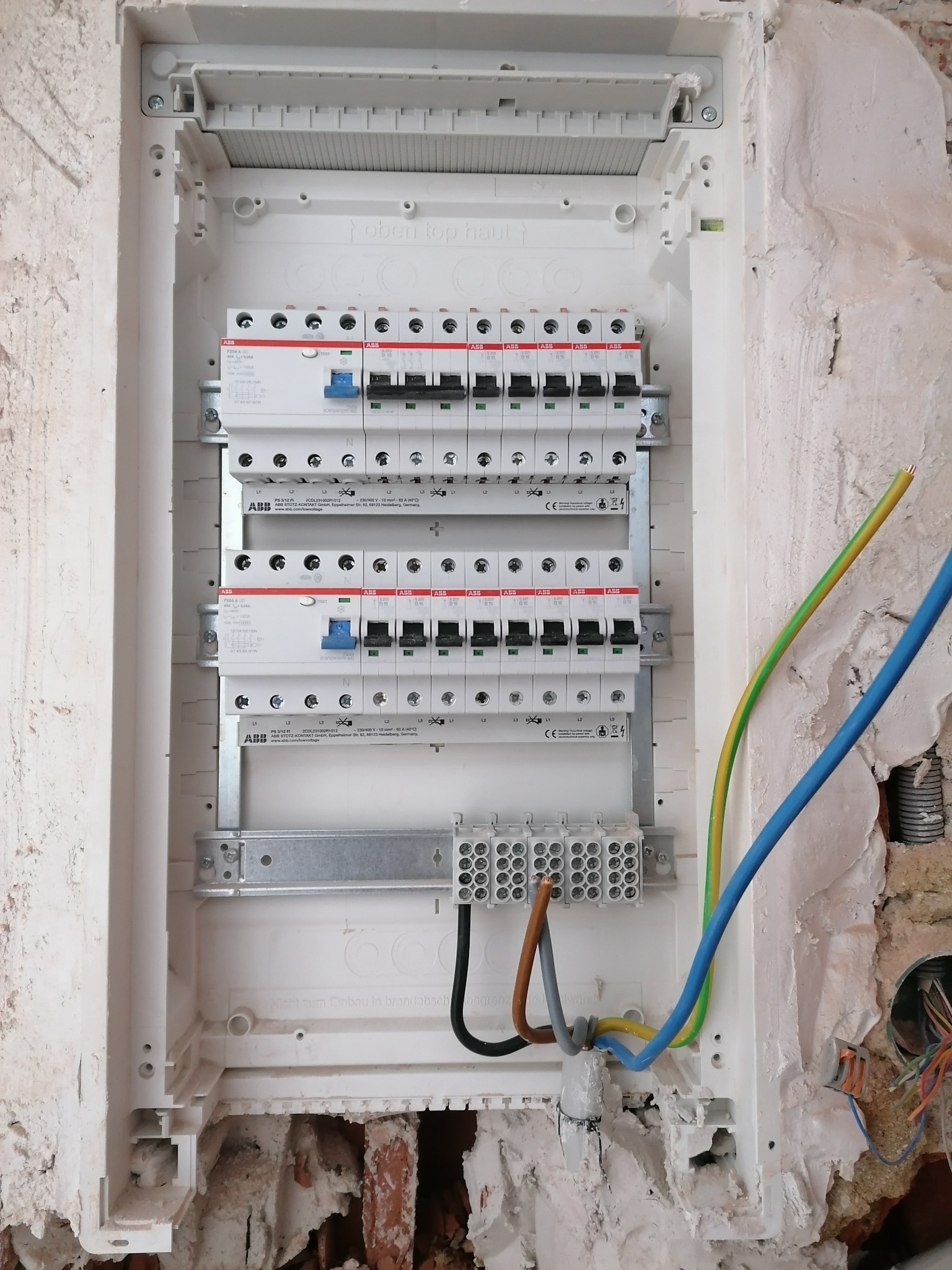 Rozdzielnica elektryczna ABB w trakcie montażu w surowej ścianie, widoczne przewody zasilające o różnych kolorach, ślady zaprawy murarskiej i niedokończone wykończenie.