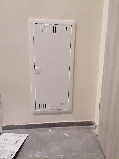 Biała, wentylowana skrzynka rewizyjna z okrągłym zamkiem, wbudowana w ścianę pomalowaną na kremowo, widoczna na tle szarej podłogi i fragmentu drzwi.