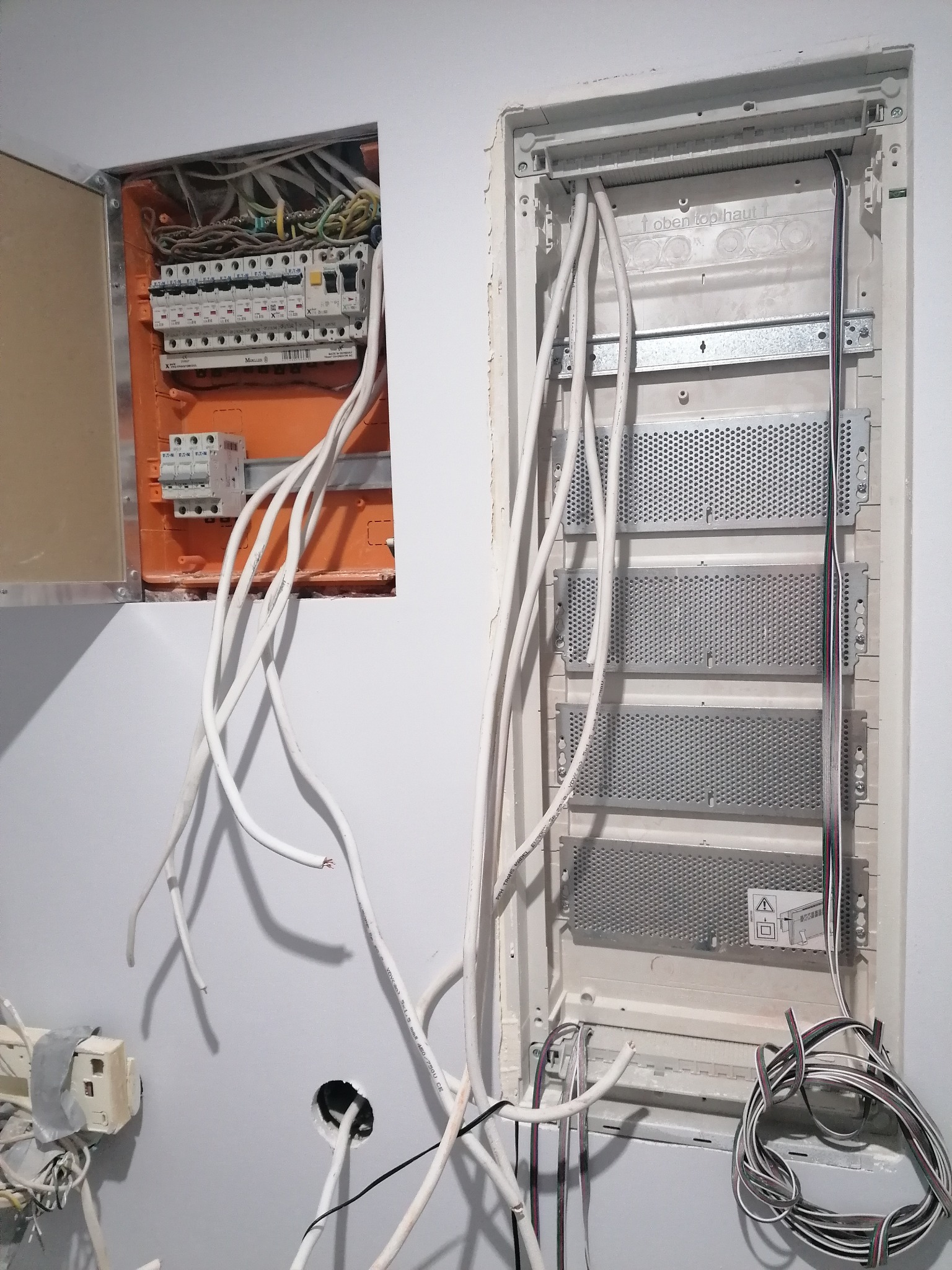 Otwarta pomarańczowa skrzynka rozdzielcza z bezpiecznikami i przewodami elektrycznymi obok białej wnęki z perforowanymi panelami i zwisającymi kablami, widoczne także gniazdko elektryczne zaklejone...