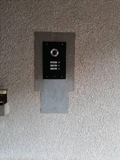 Czarny panel domofonu z trzema przyciskami i kamerą, zamontowany na szarej, chropowatej ścianie. Widoczny ślad po poprzedniej instalacji.