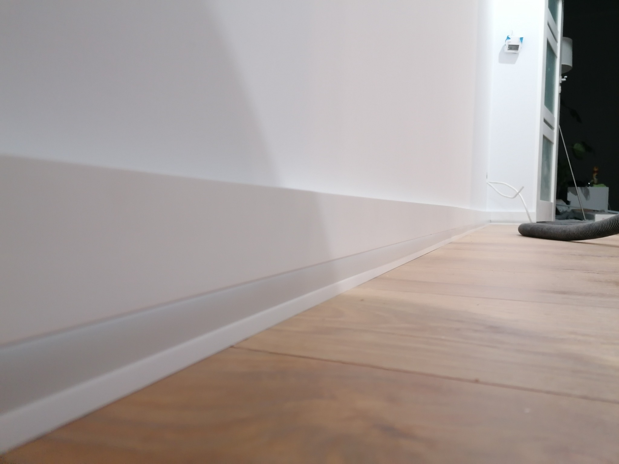 Nowoczesne białe listwy przypodłogowe zamontowane przy ścianie na drewnianej podłodze, widoczny fragment węża od odkurzacza w tle.