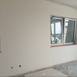 Agnieszka Jarosz Mama Na Sterydach - Biała ściana z dwoma oknami, jedno otwarte, drugie zasłonięte folią. Widoczne ślady po gniazdkach elektrycznych. Podłoga zabezpieczona folią malarską.
